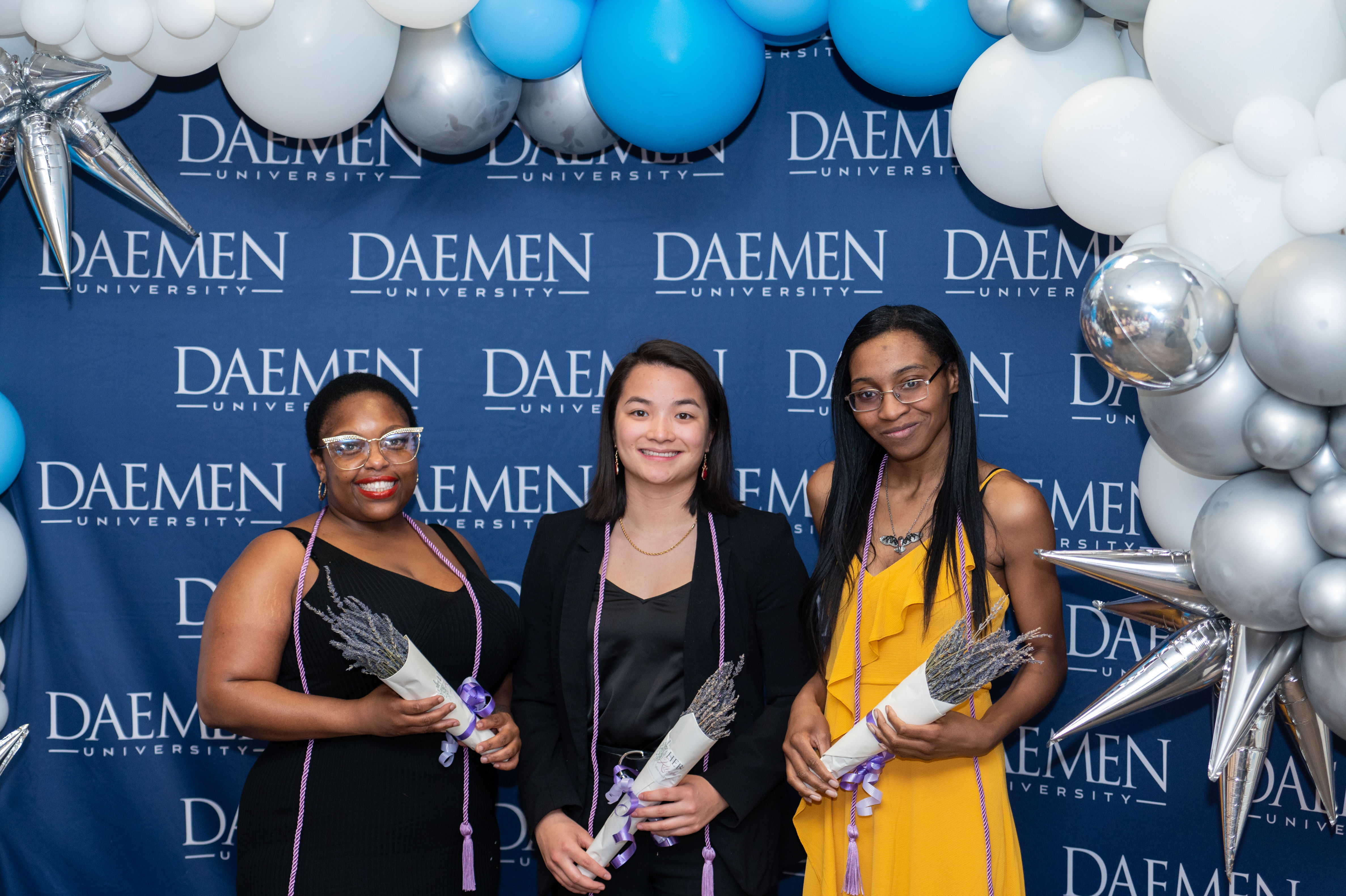 Graduates who have give approval of photo sharing standing in front of Daemen backdrop holding bouquets of lavender and wearing lavender honor ropes