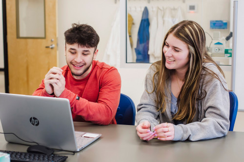 Two students sitting at a table looking at a laptop smiling