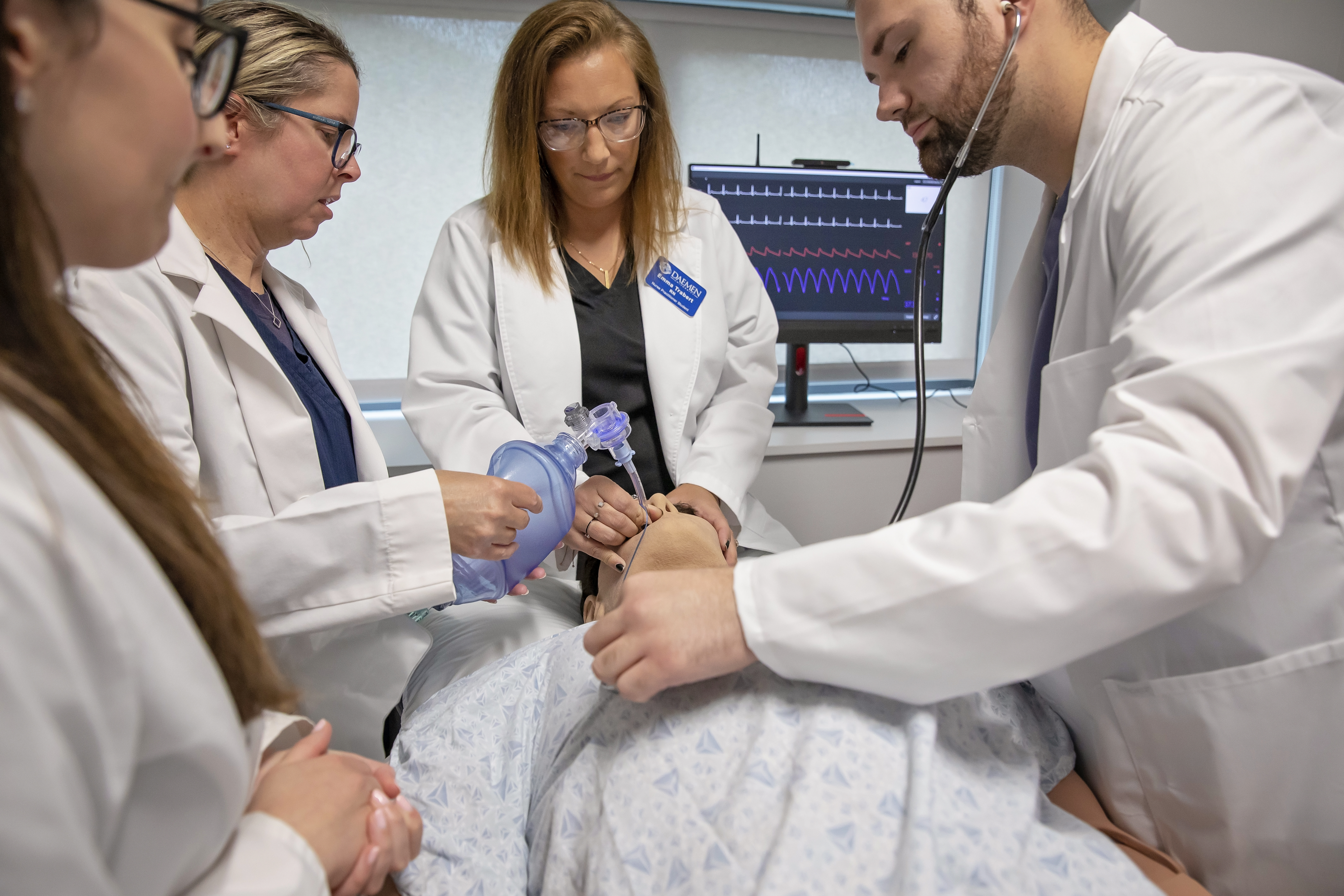 Image of professor with nursing students all wearing white coats performing medical treatment on a manikin