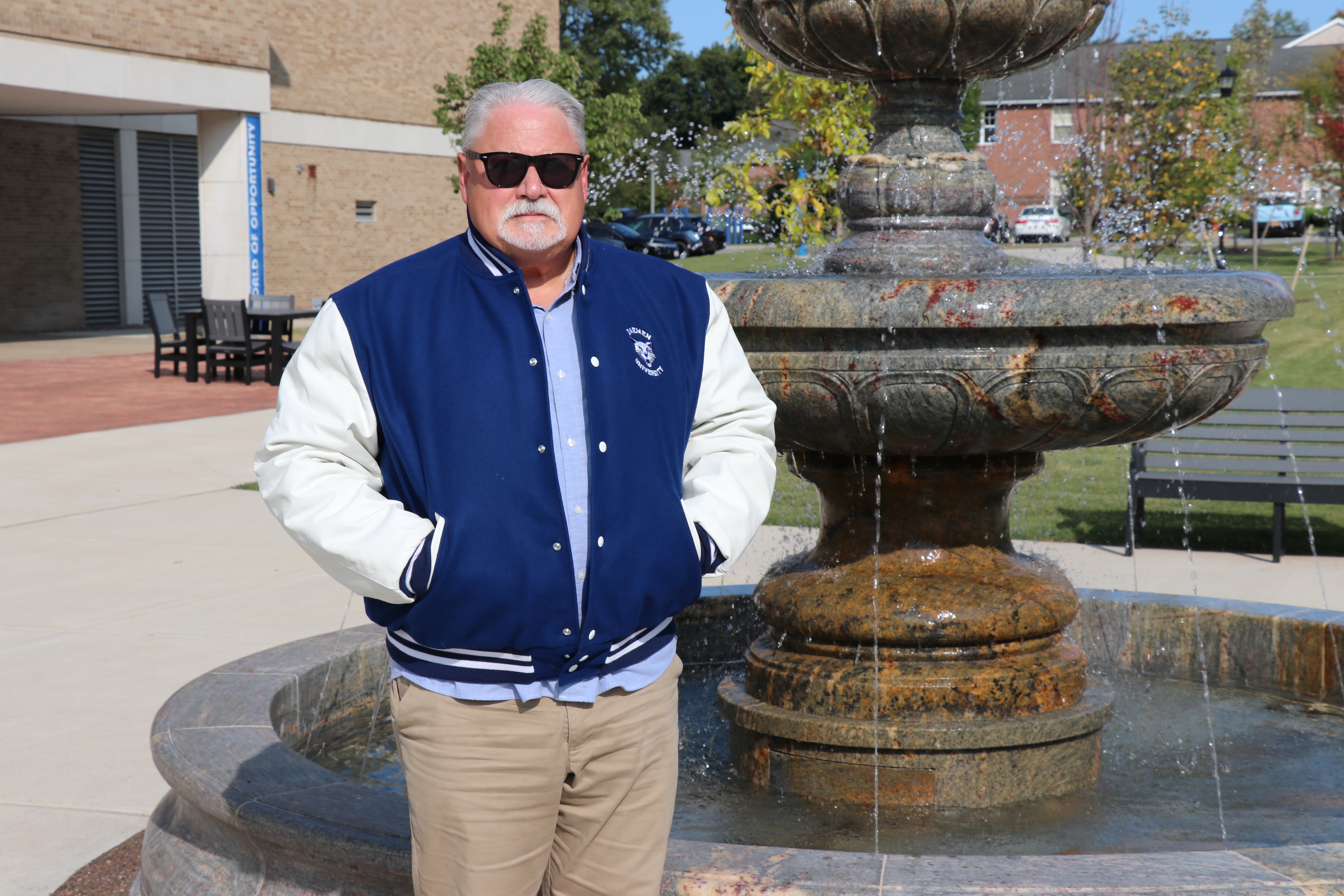 Dr. Michael Brogan wearing a varisty jacket standing in front of the Daemen fountain