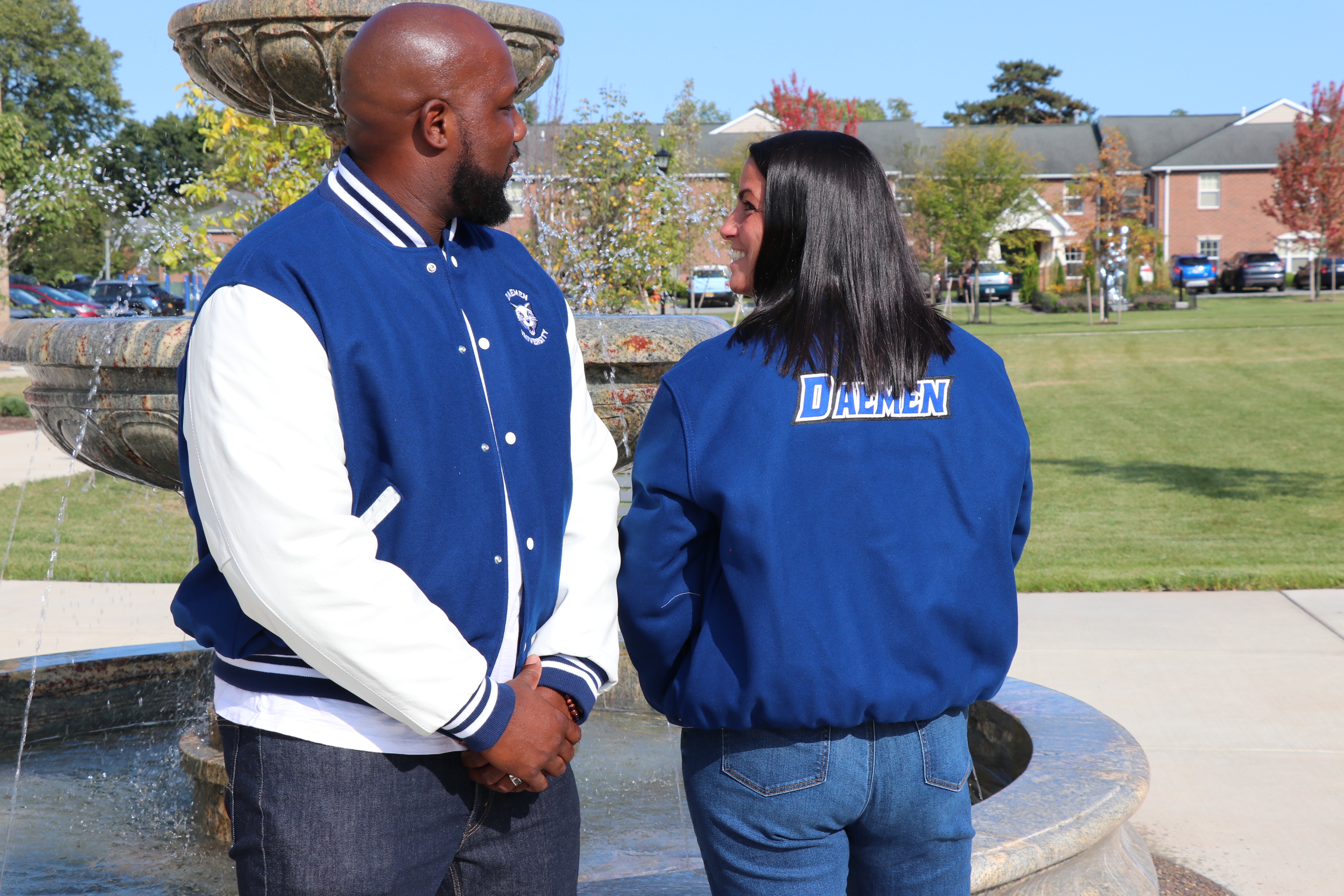 David and Bridget Burke wearing varisty jackets standing in front of the Daemen fountain