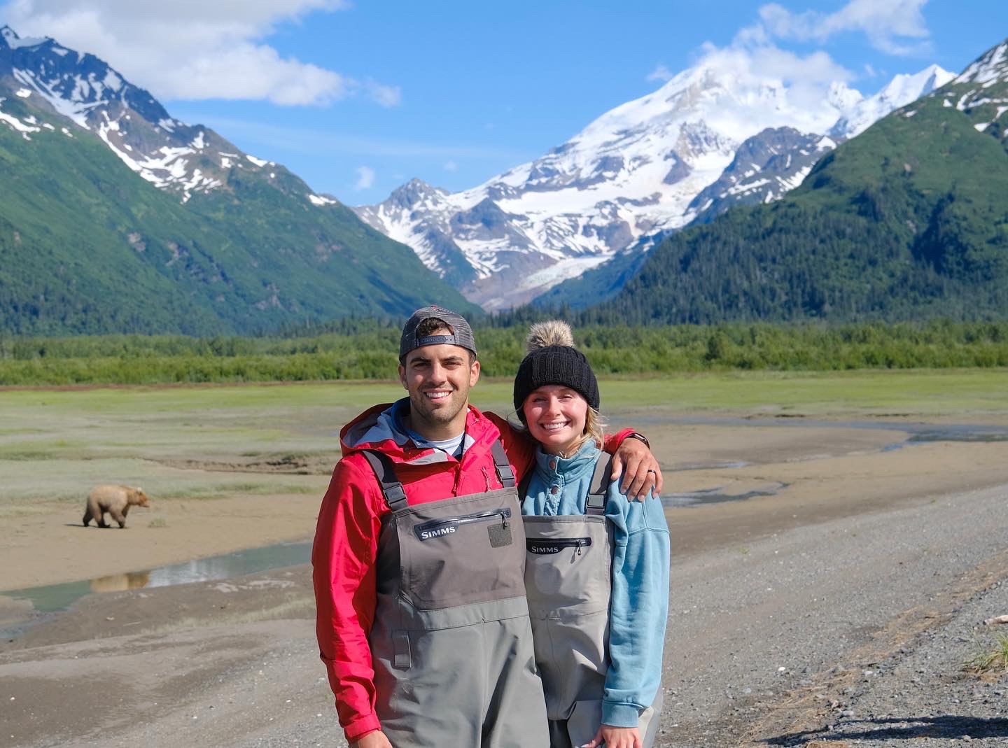 Tessa Wisniewski‘17, ‘19 and her husband standing on a shoreline with a bear and mountains behind them