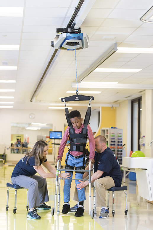 African American child wearing harness working with two therapists on walking rehab