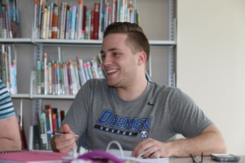 Male studentin a Daemen t-shirt smiling sitting in front of a bookshelf talking to others