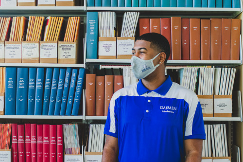 Male Daemen student wearing a face mask standing in font of a row of books in the RIC