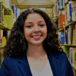 Gabby, young woman with light brown skin and shoulder lengnth curly brown hair. She is standing in an aisle of library shelves full of books, wearing a blue blazer and white top