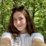 Cadence, young woman with fair colored skin, shoulder length brown hair. She has sunglasses on top of her head and is wearing a white t-shirt, standing in front of green trees 