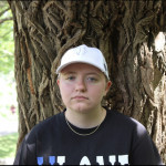 Julian, a person with short blond hair wearing a white baseball cap, stands in front of a tree wearing a dark blue sweatshirt. He has blue eyes and fair skin.