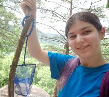 Female with long dark hair in a pony tail holding a small net with an isopod inside with a wooded background.